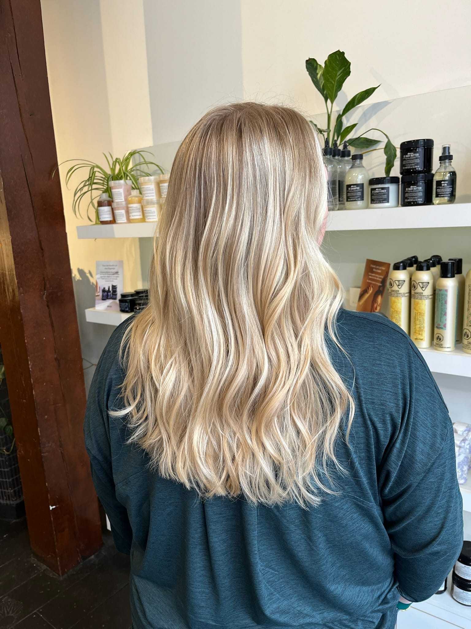 Blonde-haired person in a salon, standing before shelves of hair products.