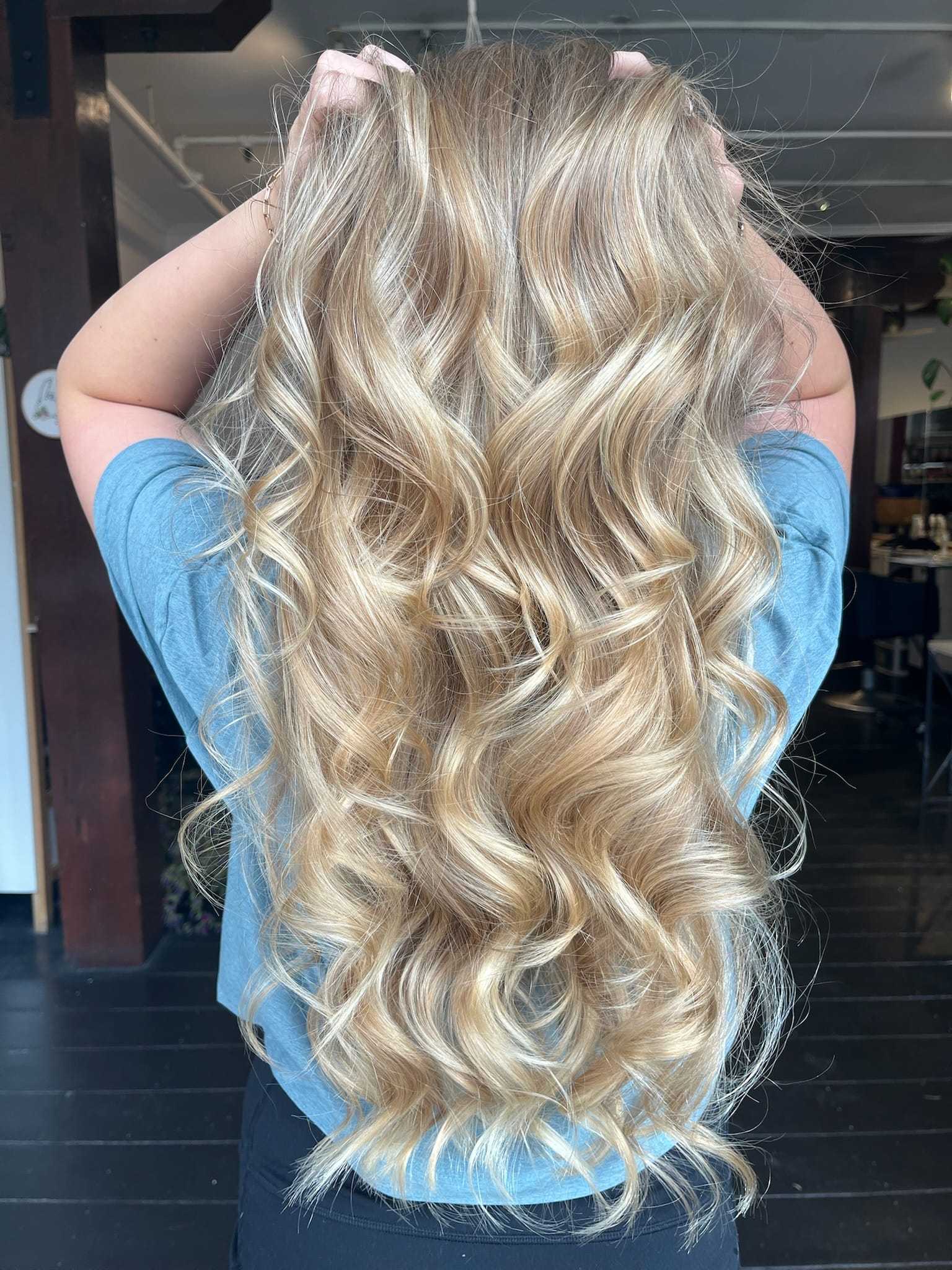 Long, wavy blonde hair flowing down a person's back, hands in hair, blue shirt.