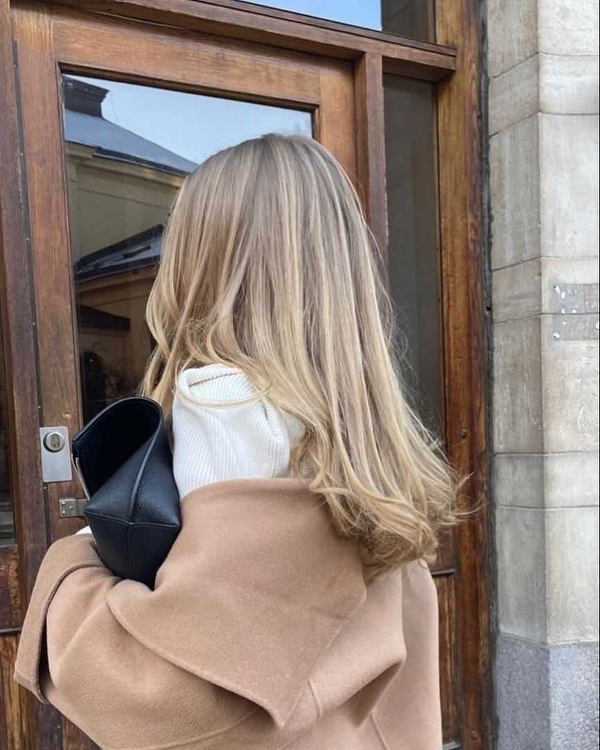 Woman with long blonde hair holding a black bag, standing in front of a wooden door.